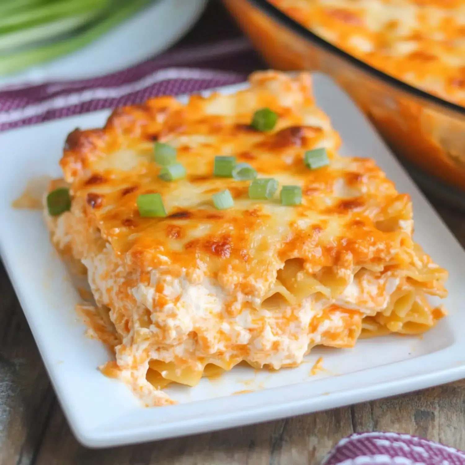 A rich, creamy lasagna slice garnished with green onions on a white plate, with the full dish in the background.
