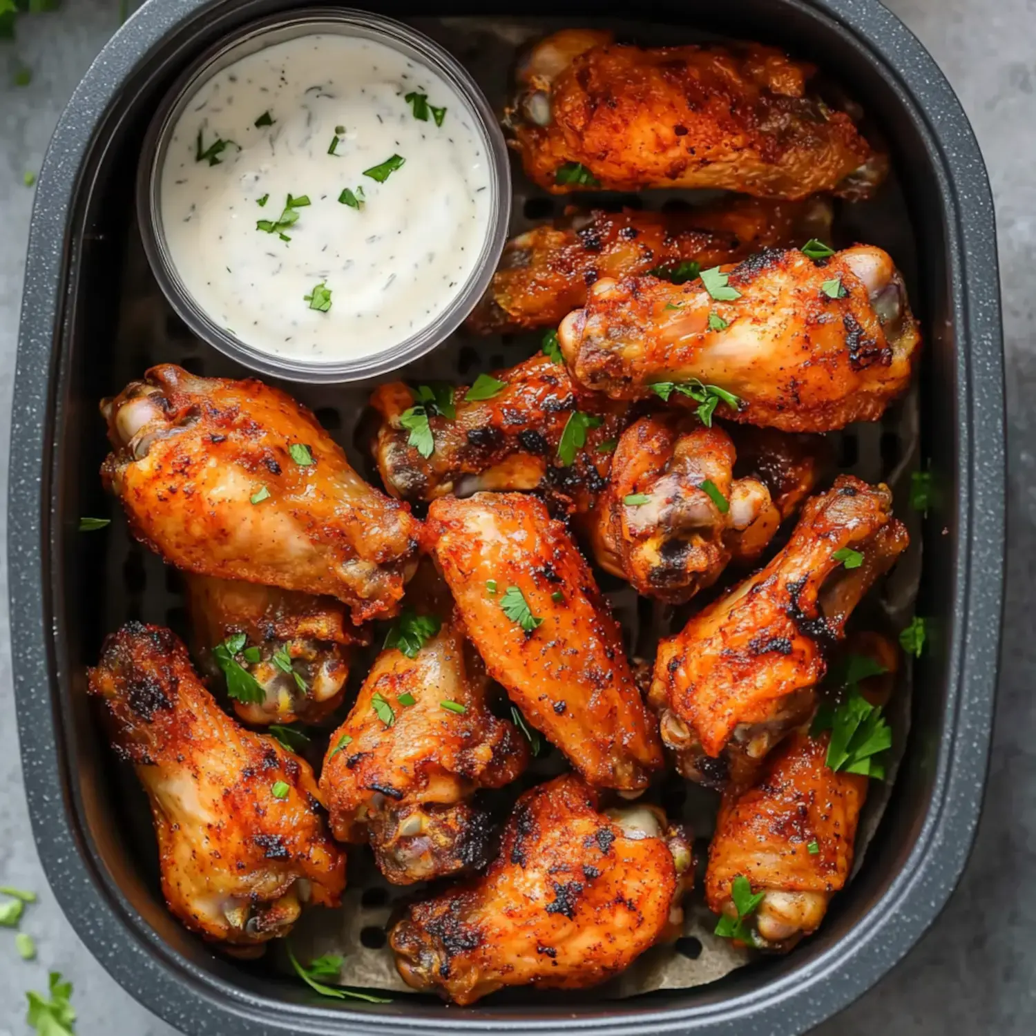 A plate of golden, crispy chicken wings sprinkled with parsley, served next to a cup of dipping sauce.