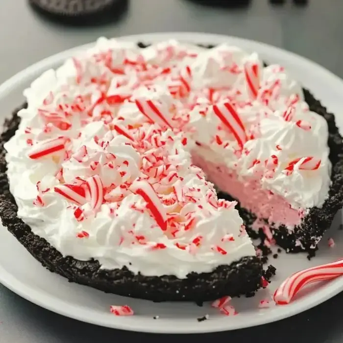 A creamy peppermint pie with a chocolate crust, covered in whipped cream and candy cane bits, with one slice missing.