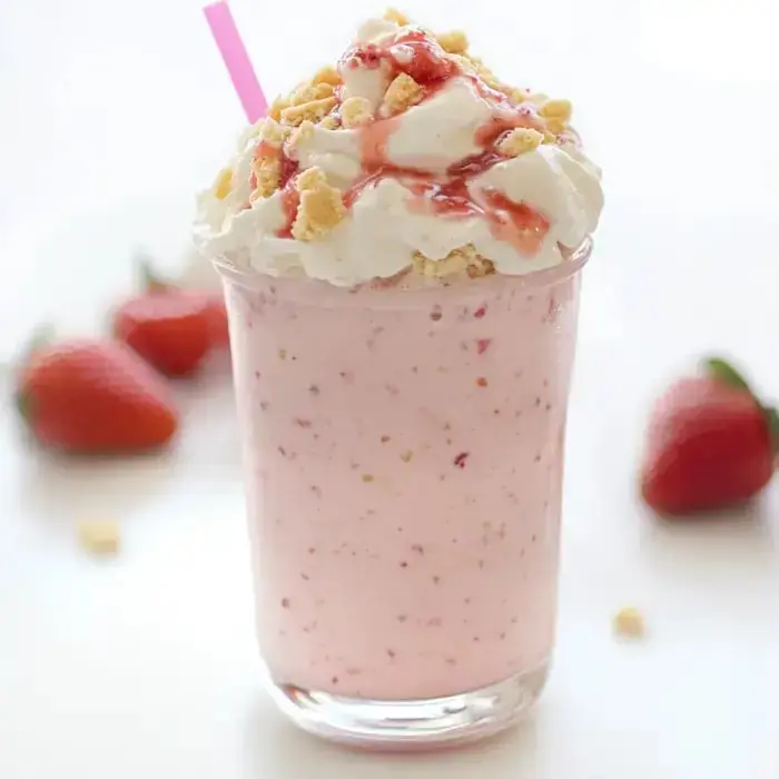 A creamy pink milkshake topped with whipped cream, cookie crumbs, and strawberry sauce, with fresh strawberries on the side.