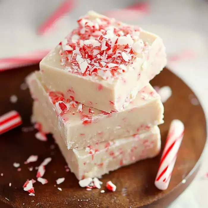 Blocks of white peppermint fudge with crushed candy canes on a wooden board next to small candy canes.