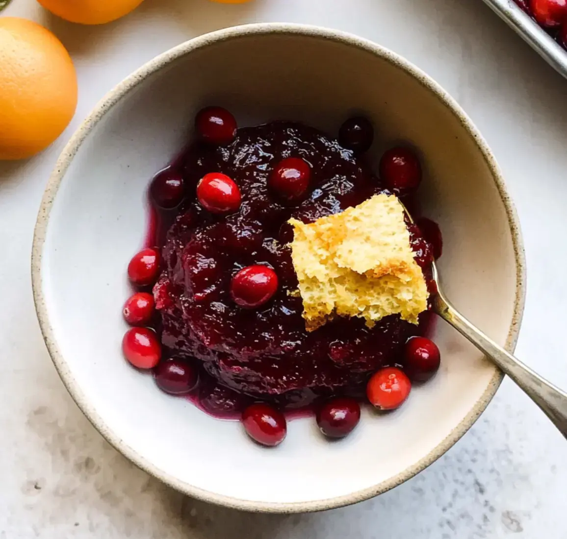 A bowl of cranberry sauce garnished with fresh cranberries and cornbread, with oranges beside it.
