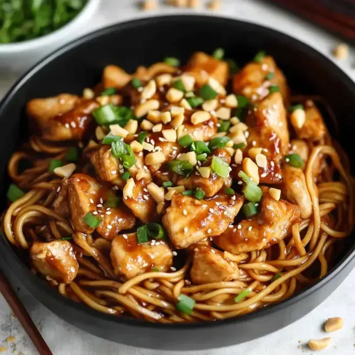Close-up of black bowl packed with noodles, chicken bits, chopped green onions, crushed peanuts, and sesame seeds in a delicious glossy sauce.