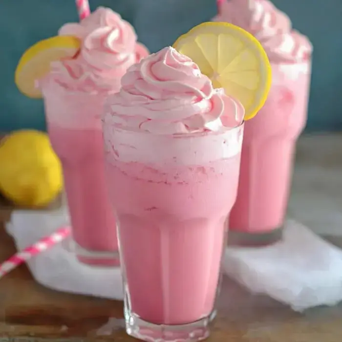 Three frothy pink drinks in tall glasses, topped with whipped cream, lemon wedges, and paper straws on the side.