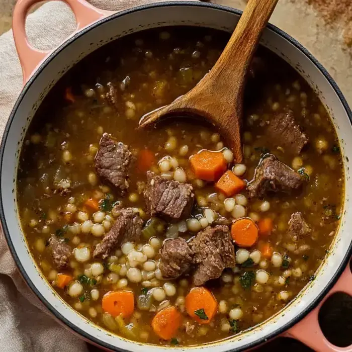 A wooden spoon stirring a hearty pot of beef soup with diced carrots and pearl couscous.