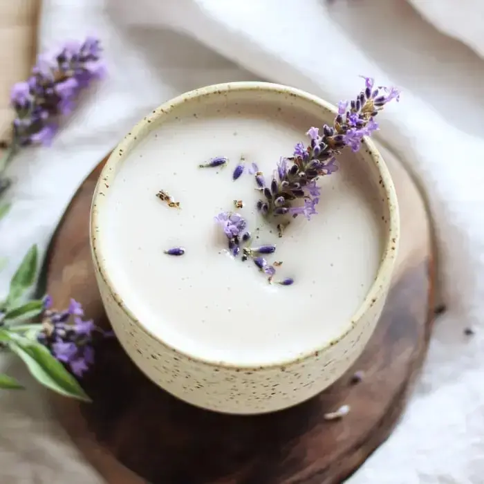 A close-up of a creamy drink topped with fresh lavender, sitting in a speckled mug on a wood table.