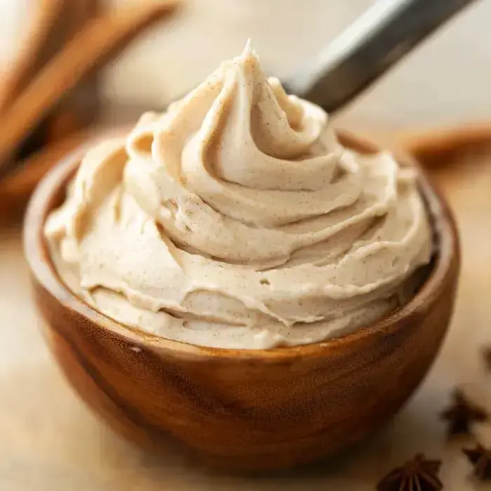 Swirls of creamy, chai-colored frosting in a wooden bowl with cinnamon sticks on the side.