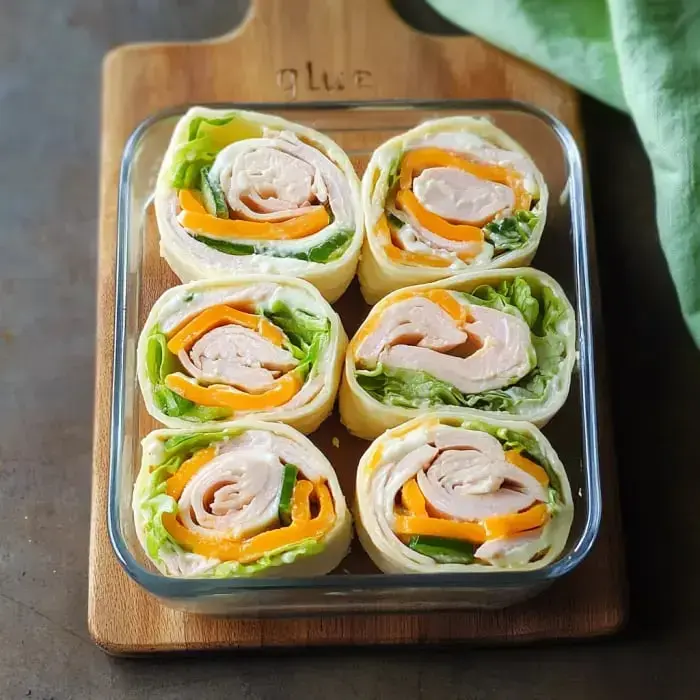 Six rolled tortilla snacks packed with lettuce, turkey, and cheddar, all sitting neatly in a glass pan.