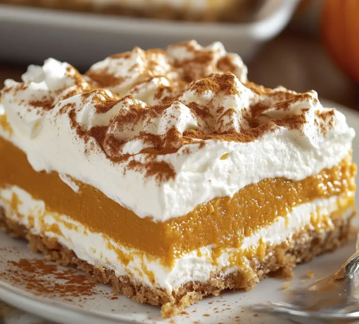 A close-up view of a pumpkin dessert slice stacked with fluffy whipped cream on a crumbly cookie base and sprinkled with cinnamon.