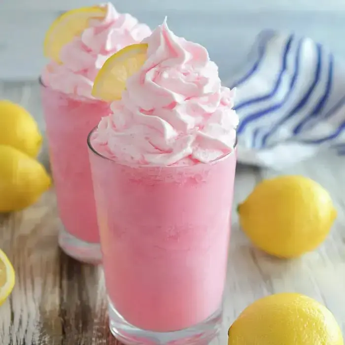 A pair of creamy, frothy pink lemon drinks topped with whipped cream and lemon slices sit next to fresh lemons on a wooden board.