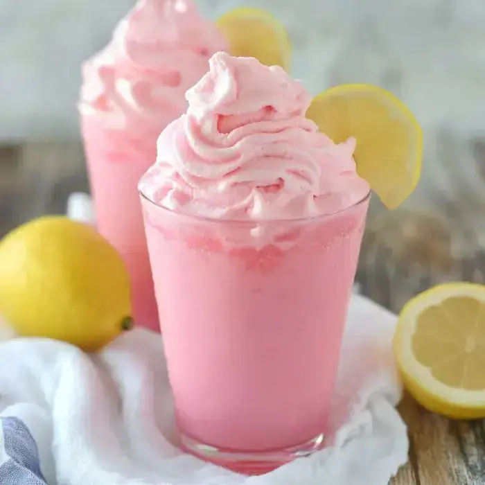 Pink whipped lemonades are dressed up with lemon wedges and fresh lemons sit nearby on a white cloth.