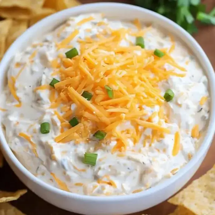 A bowl packed with creamy dip, topped with cheddar and green onions and some chips on the side.