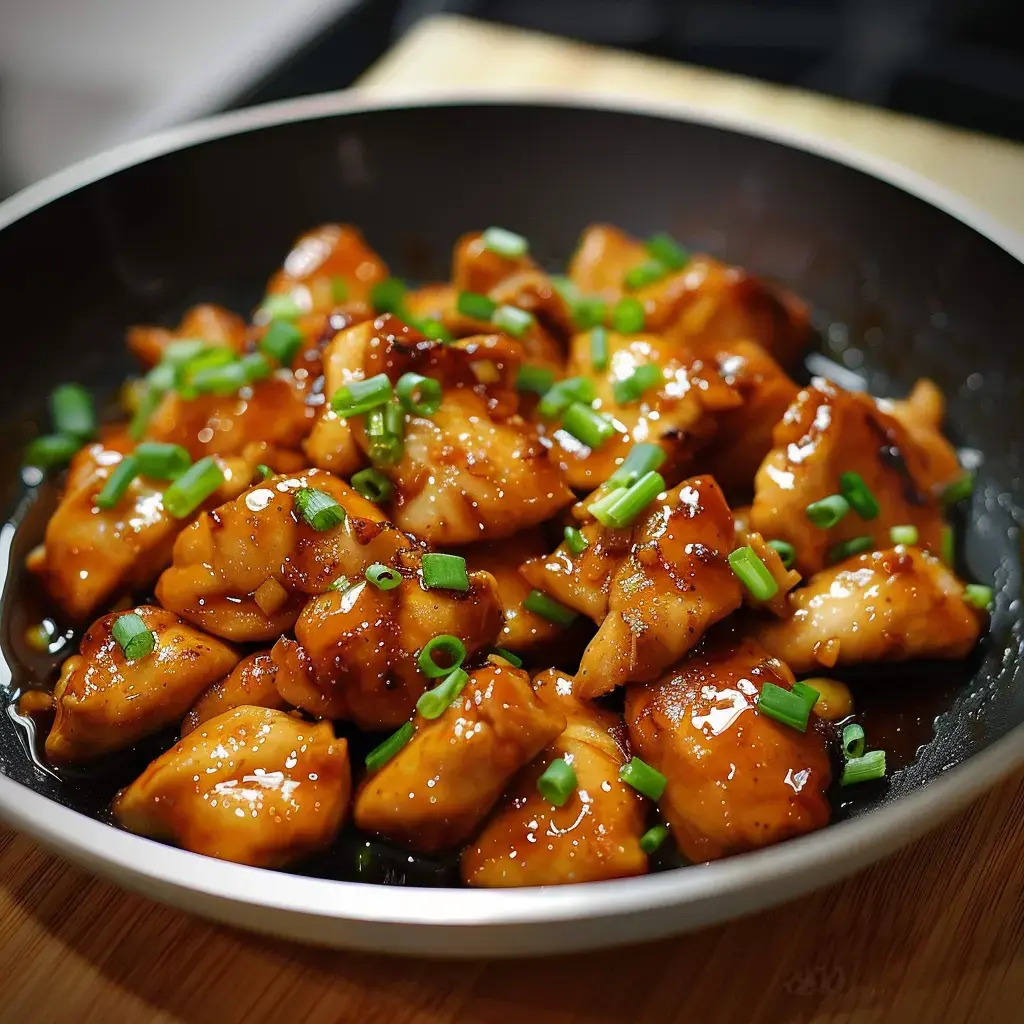 A bowl of glazed chicken with chopped chives on top, served in a black dish.