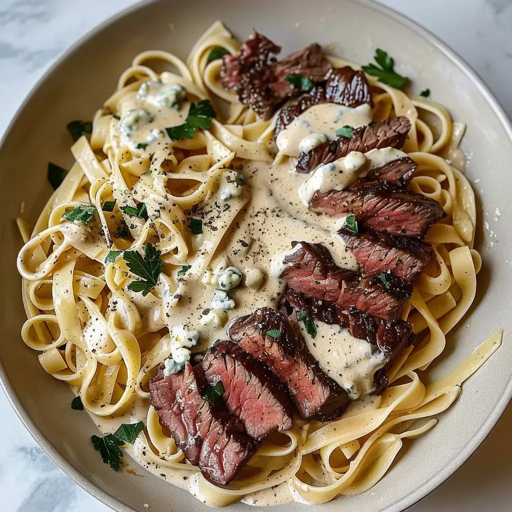 A plate of fettuccine pasta topped with slices of steak and creamy sauce, garnished with parsley and blue cheese.