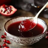 A spoon tugs at glossy, ruby-red pomegranate jelly from a detailed bowl, pomegranate seeds scattered nearby.