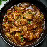 Pieces of tender chicken coated in a savory sauce, topped with fresh herbs, served in a bowl.