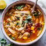 A bowl of steamy soup filled with chicken, corn, tomatoes, and fresh cilantro, served with lime and green pepper on the side.