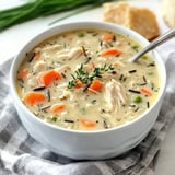 A bowl of creamy chicken and wild rice soup with carrots, peas, and fresh thyme garnish.