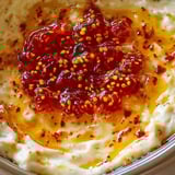 A close shot of a creamy cheesy dip topped with bright red pepper jelly and mustard seeds.