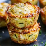 Two golden zucchini muffins stacked up with crispy edges and green herb flecks, resting on a dark surface.