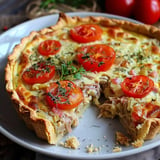 Savory tart topped with tomatoes, herbs, and chunks of meat, with a slice cut out.