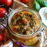 A glass jar filled with a gourmet mix of tomatoes, herbs, and spices, surrounded by fresh tomatoes and cloves of garlic.